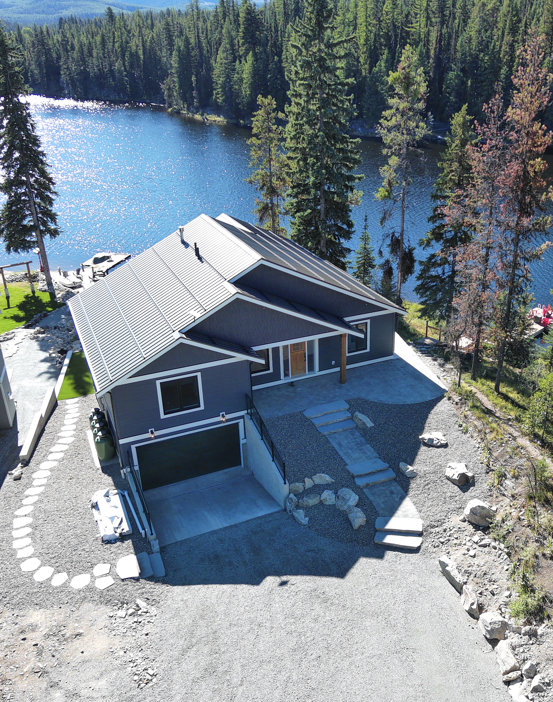 A high-resolution architectural photograph of a modern waterfront custom home in Vancouver, featuring a sleek metal roof, dark siding, and professional landscaping overlooking a lake and forest.